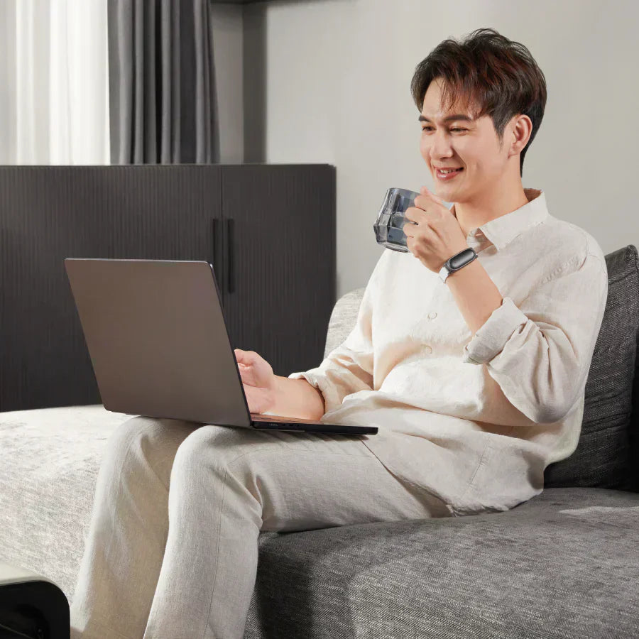 A man smiling while working on a laptop and drinking from a mug, sitting comfortably on a gray sofa.
