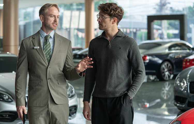 A car salesman wearing a silver Plaud NotePin on his lapel explains features to a customer in a showroom.