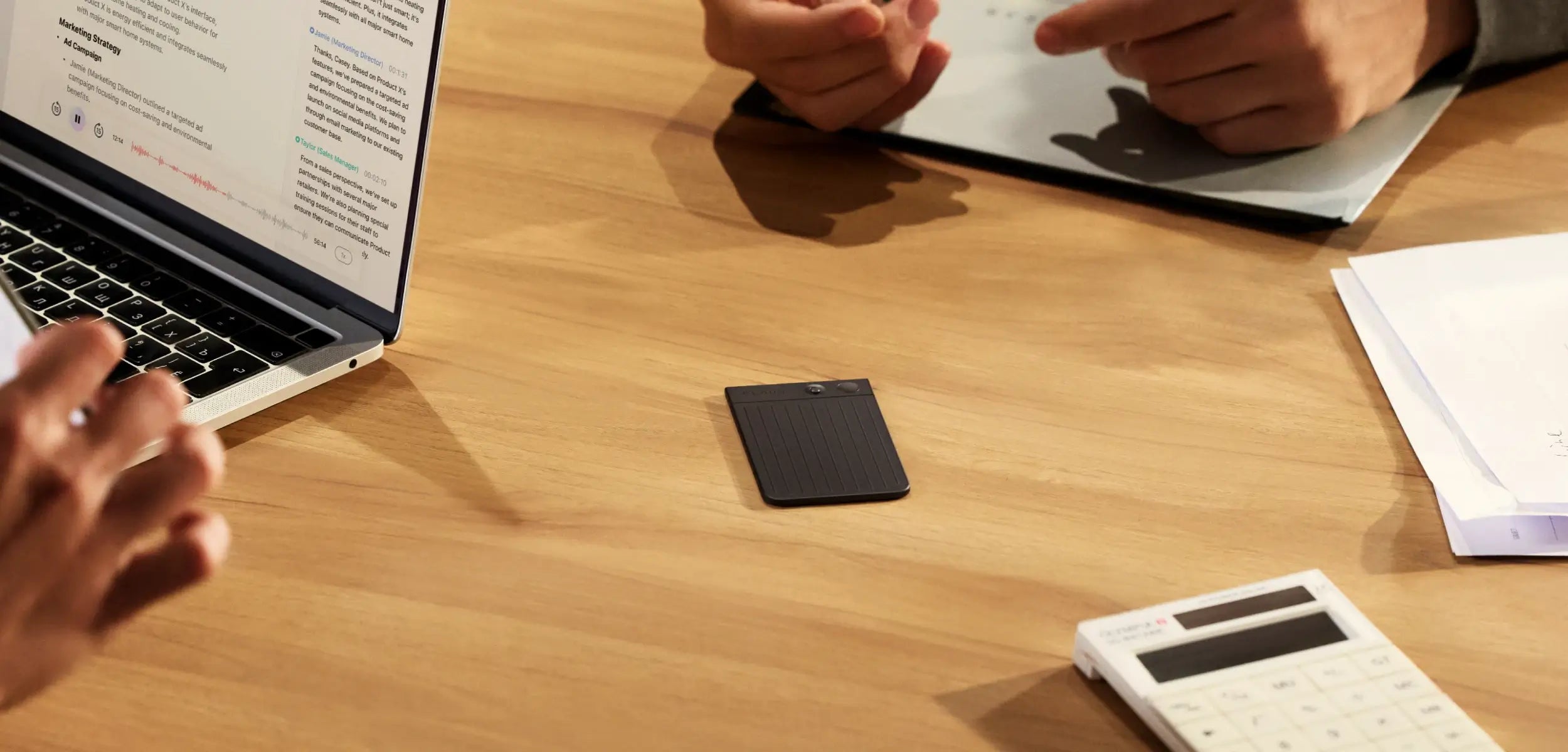 Black Plaud Note on a wooden table during a meeting, with a laptop, hands, and papers nearby.
