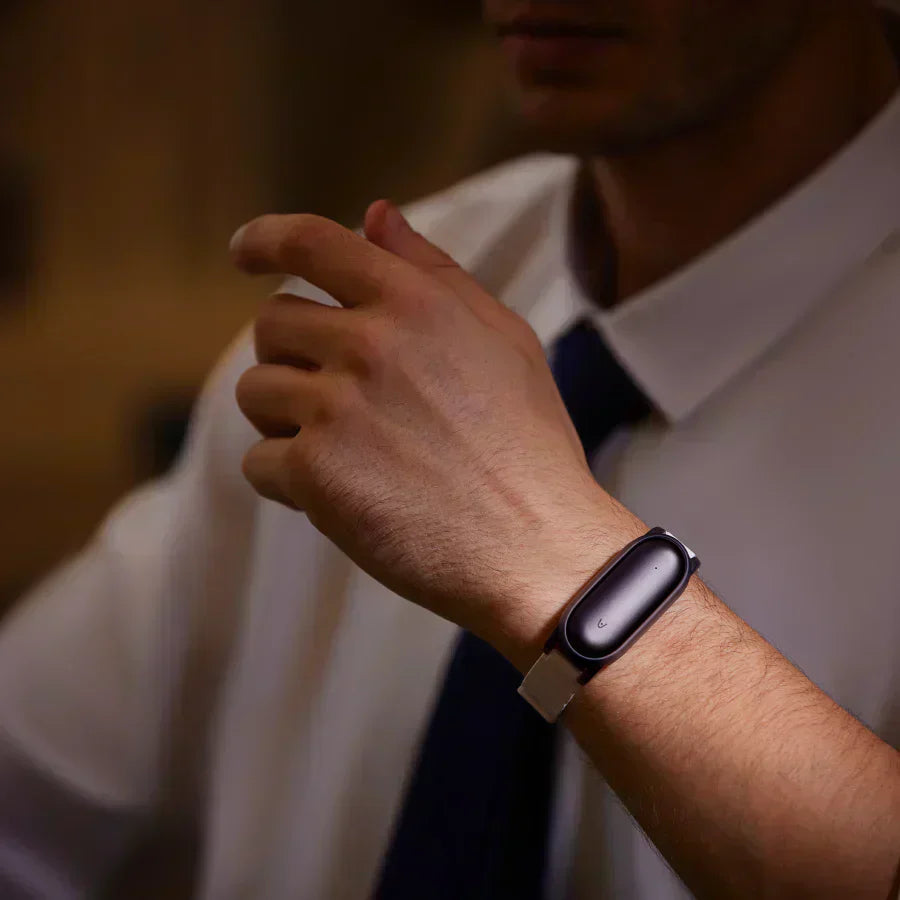 A person wearing a dark grey Plaud NotePin on a light grey wristband, with a white shirt and dark tie.