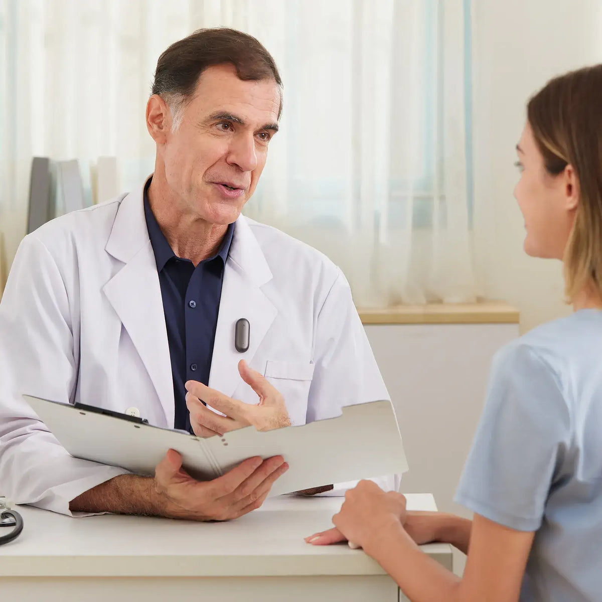 A doctor wearing a dark gray Plaud NotePin on his white lab coat, talking to a patient during a consultation.
