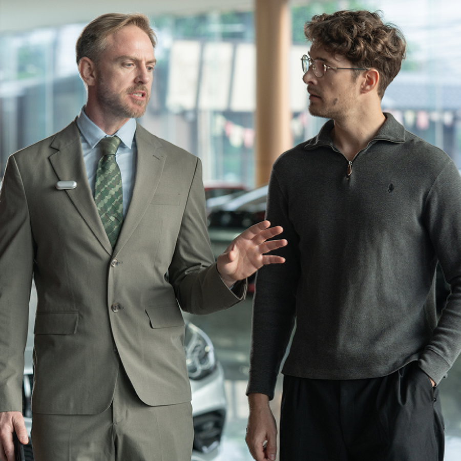 A salesman wearing a silver Plaud NotePin on his lapel explains features to a customer in a car dealership.