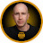 Bald man in a dark t-shirt with a gold emblem, framed in a gold circle.