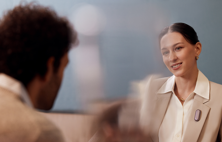 Woman in a blazer wearing a muted purple Plaud NotePin S, smiling at a person across from her in a professional setting.