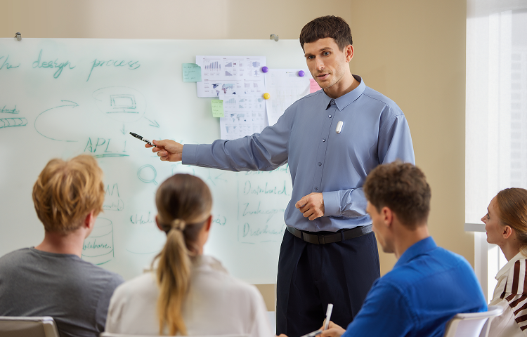 A presenter wearing a white Plaud NotePin on his shirt points to a whiteboard during a business meeting.