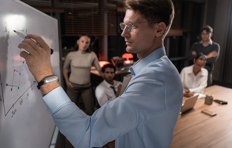 Man presenting a graph on a whiteboard, wearing a silver Plaud NotePin on a black wristband during a meeting.