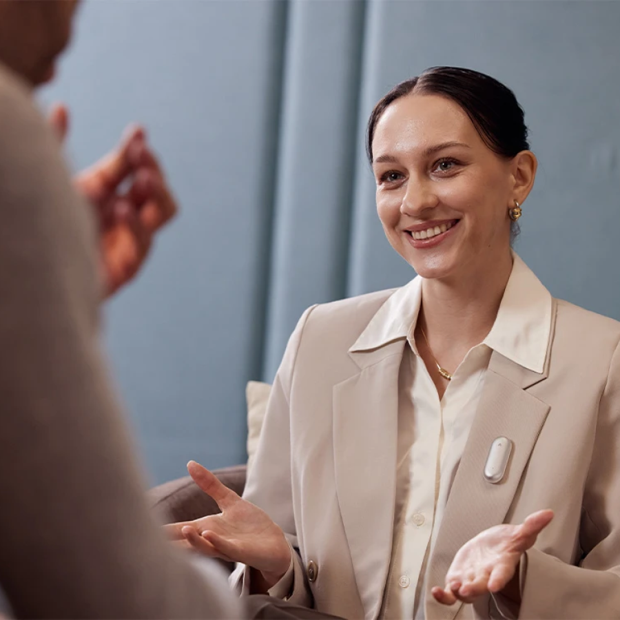 Smiling woman in a blazer wearing a white Plaud NotePin, gesturing while talking to another person.