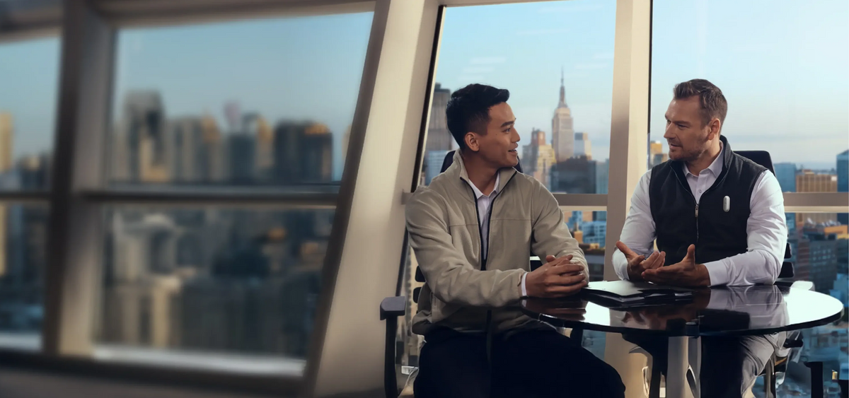 Two men in a modern office with a city skyline view, one wearing a Plaud NotePin, discussing business.