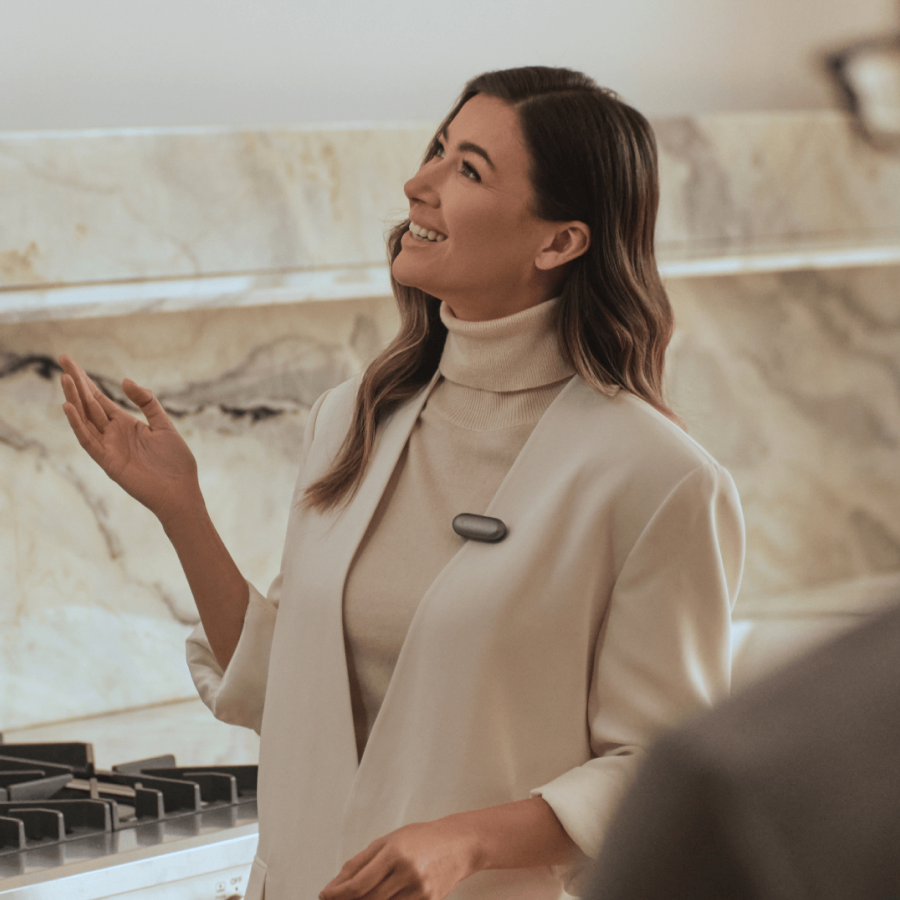 Woman wearing a dark gray Plaud NotePin on her cream blazer, smiling and gesturing.