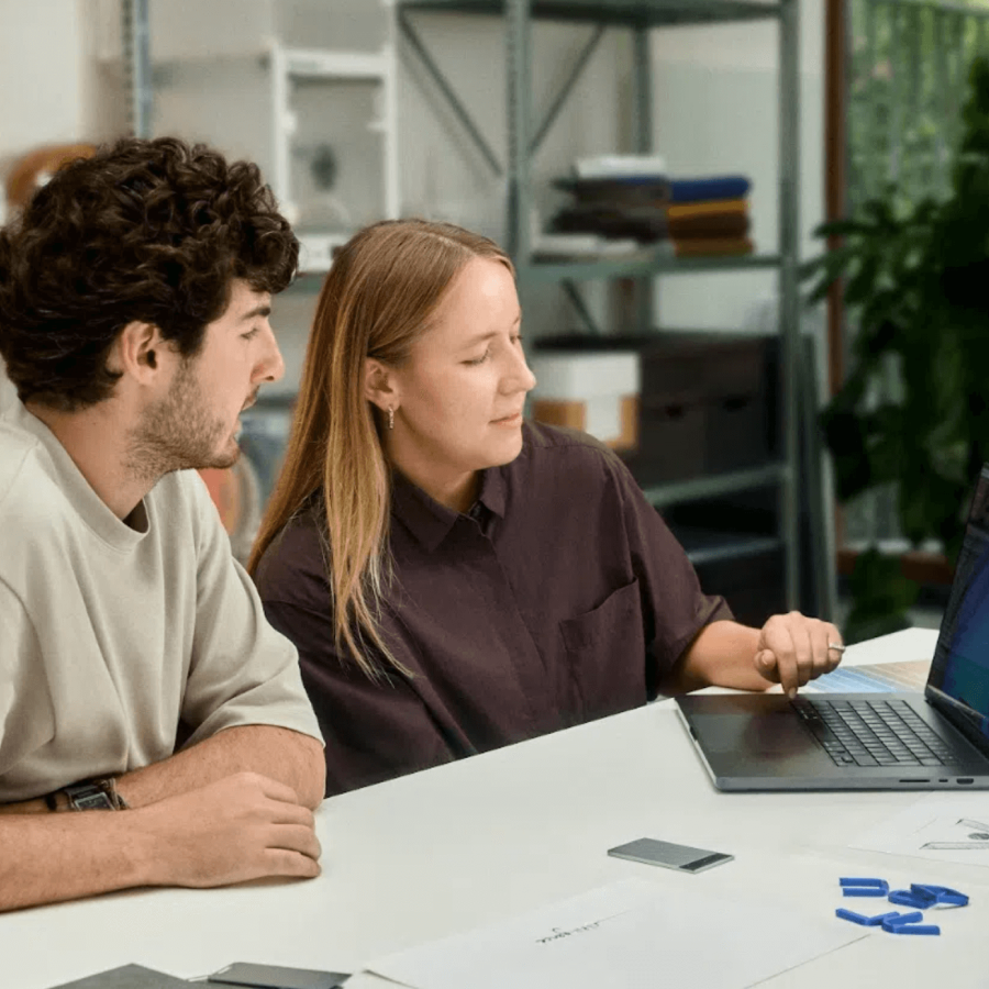 Two colleagues collaborating at a white table, looking at a laptop screen. A smartphone and papers are on the table.