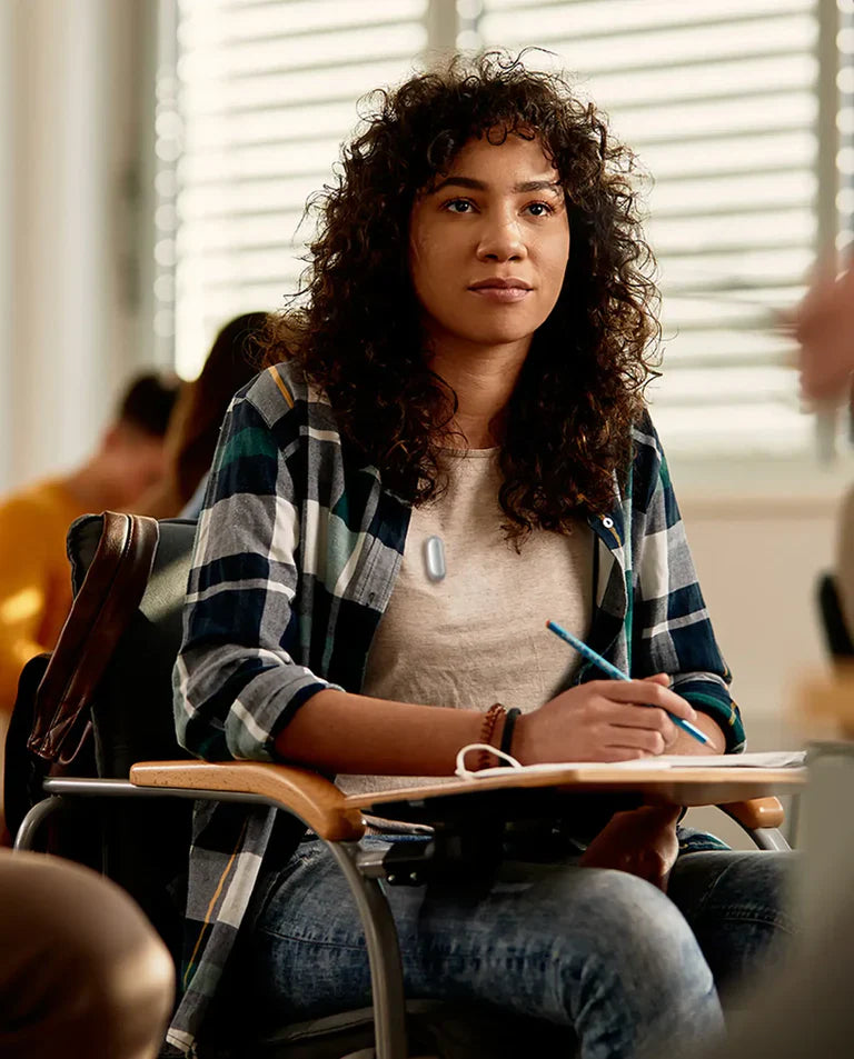 Young woman in a classroom, wearing a silver Plaud NotePin, attentively taking notes.