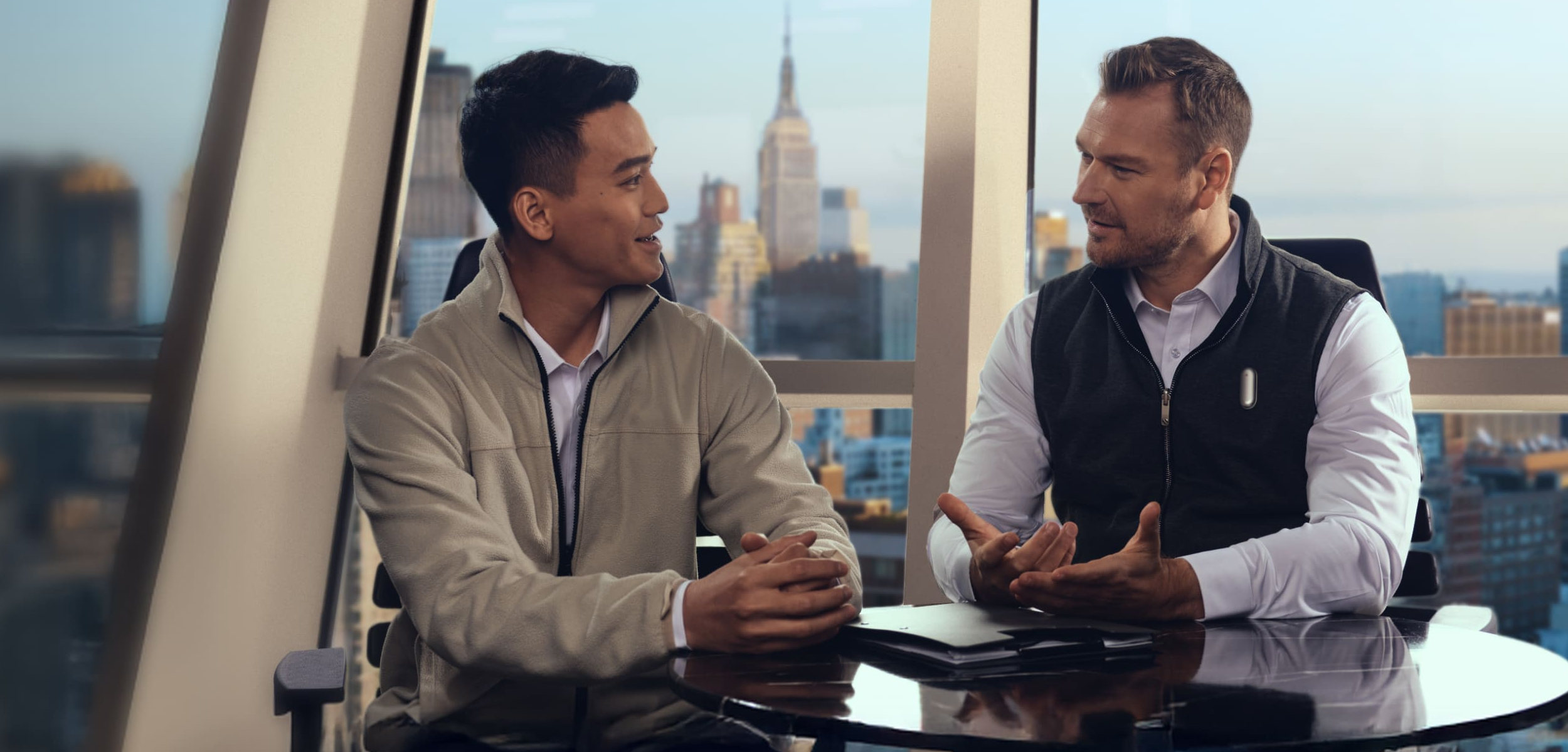 Two men in an office with a city view, one wearing a Plaud NotePin, engaged in conversation.