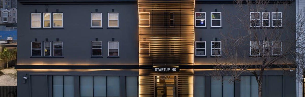 Modern dark gray building with illuminated wooden facade and 'STARTUP HQ' sign at dusk.