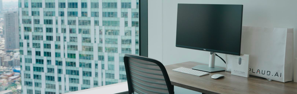 Modern office desk with computer setup and two white Plaud.ai branded bags, overlooking a city skyline from a high-rise windo
