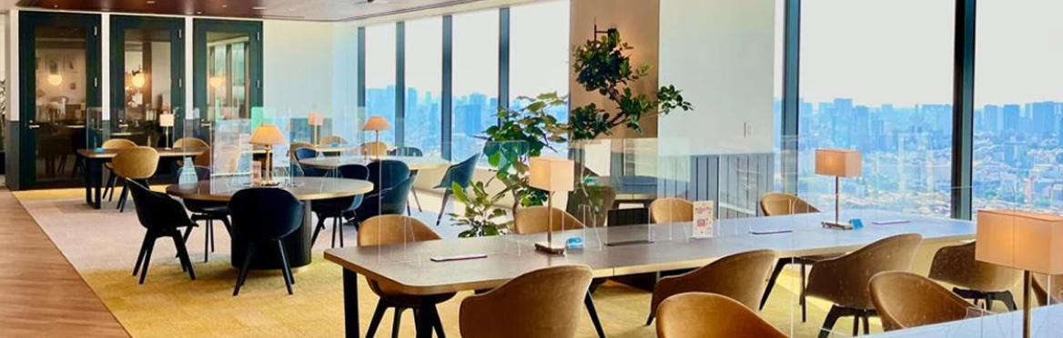 A modern, bright co-working space with large windows overlooking a city, featuring various tables, chairs, and plants.