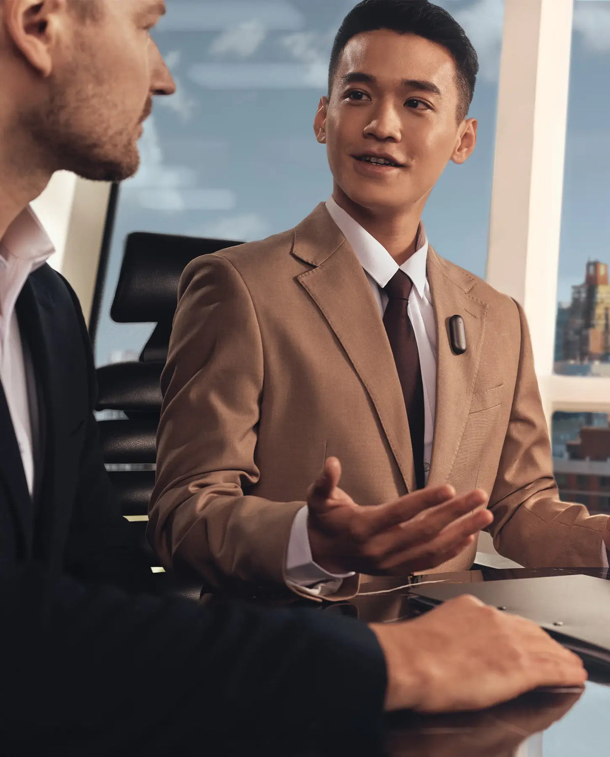 A man in a tan suit speaks during a meeting, wearing a black Plaud NotePin on his lapel.