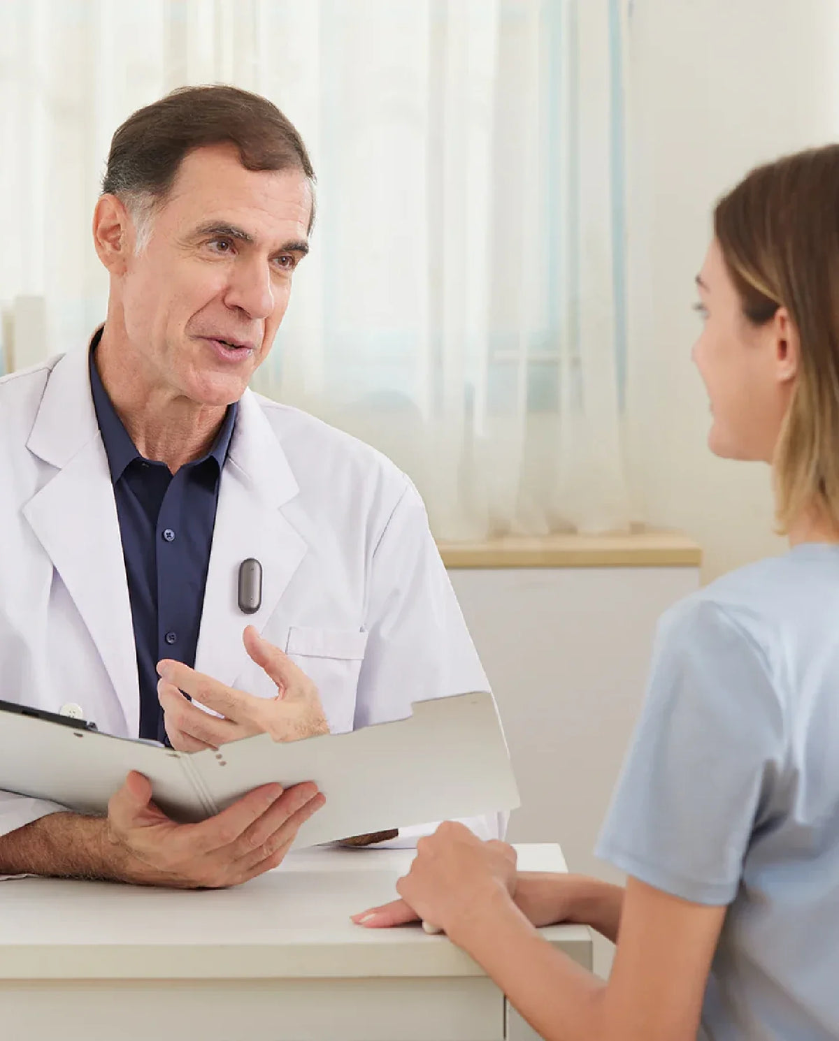 Doctor wearing a dark grey Plaud NotePin on his lab coat, talking to a patient in a medical office.