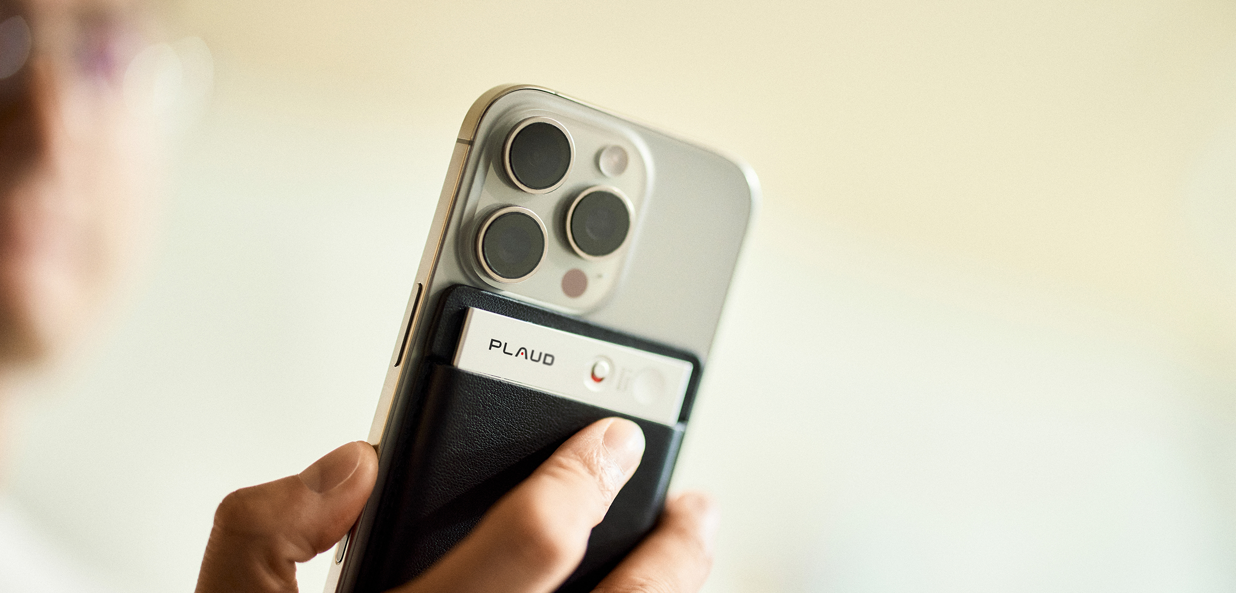 A person holds a silver smartphone with a black Plaud Note attached to its back.