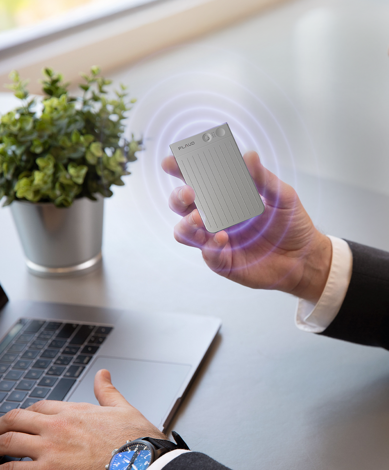 A person holds a silver Plaud Note Pro, emitting purple signal rings, above a laptop in an office setting.