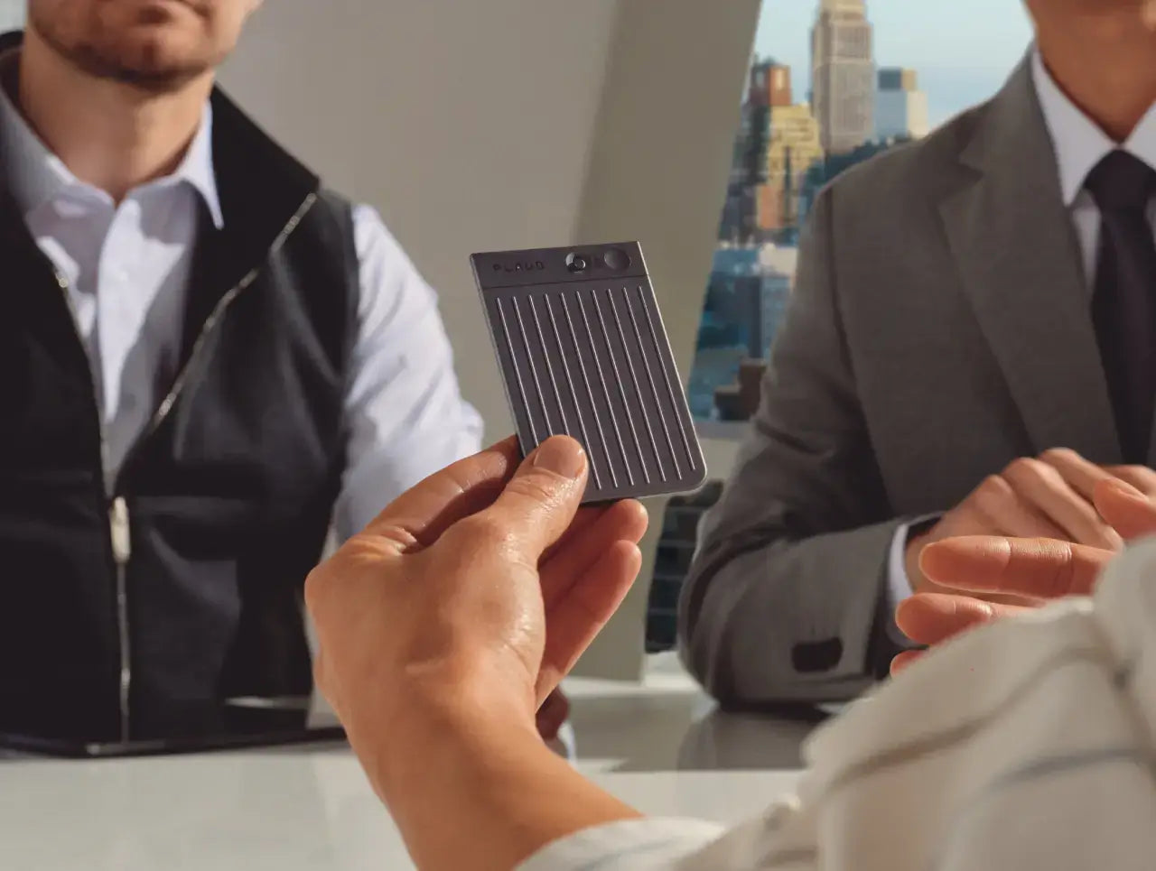 A hand holds a dark gray Plaud Note with a ribbed texture and small screen, in a business meeting setting.