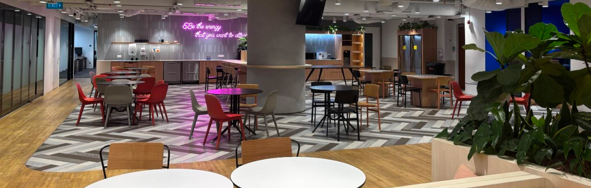 Modern office lounge with colorful chairs, tables, kitchen area, and plants, featuring a neon sign.