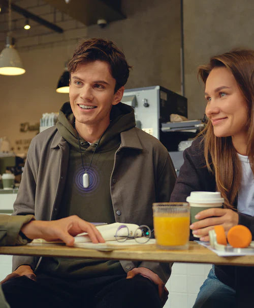 Man wearing a white Plaud NotePin on a lanyard, glowing purple, while talking with a woman in a cafe.