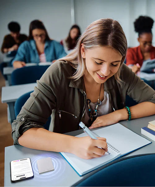 A student writes in a notebook with a silver Plaud Note and smartphone on her desk, recording a lecture in a classroom.