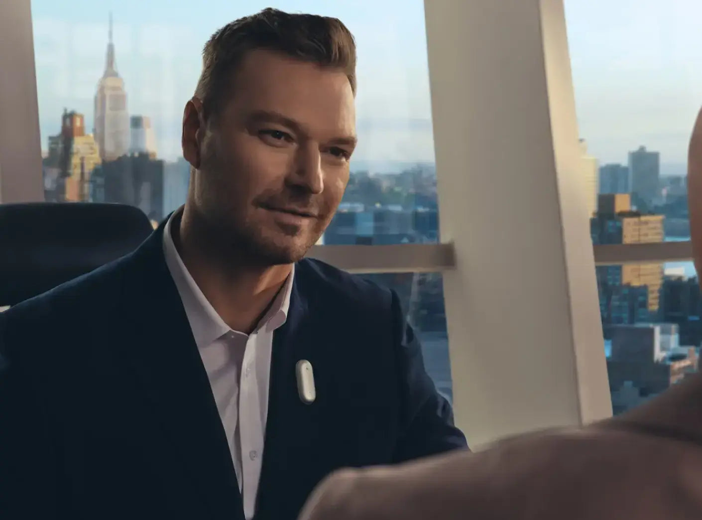Man in a suit wearing a white Plaud NotePin on his lapel, smiling in a high-rise office with a city skyline view.