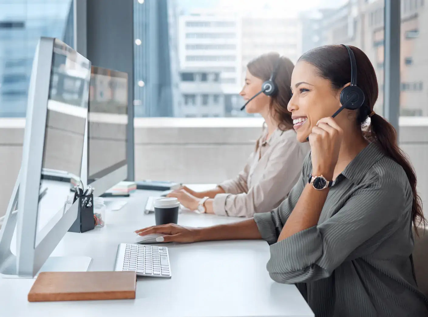 Two smiling women in headsets work at computers in a modern office, representing customer support or efficient communication.
